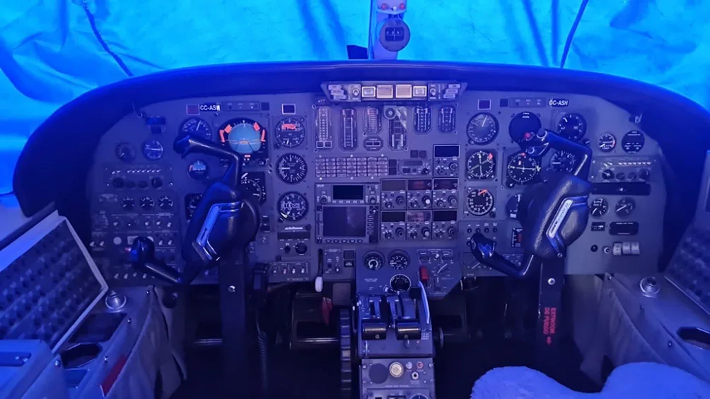 The cockpit of an aircraft with a complex instrument panel, multiple gauges, and two control yokes, illuminated by a blue light.