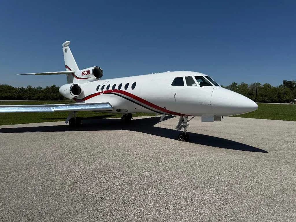A white Dassault Falcon 900EX business jet with red and black stripes on the tarmac under a clear blue sky, viewed from the front right.