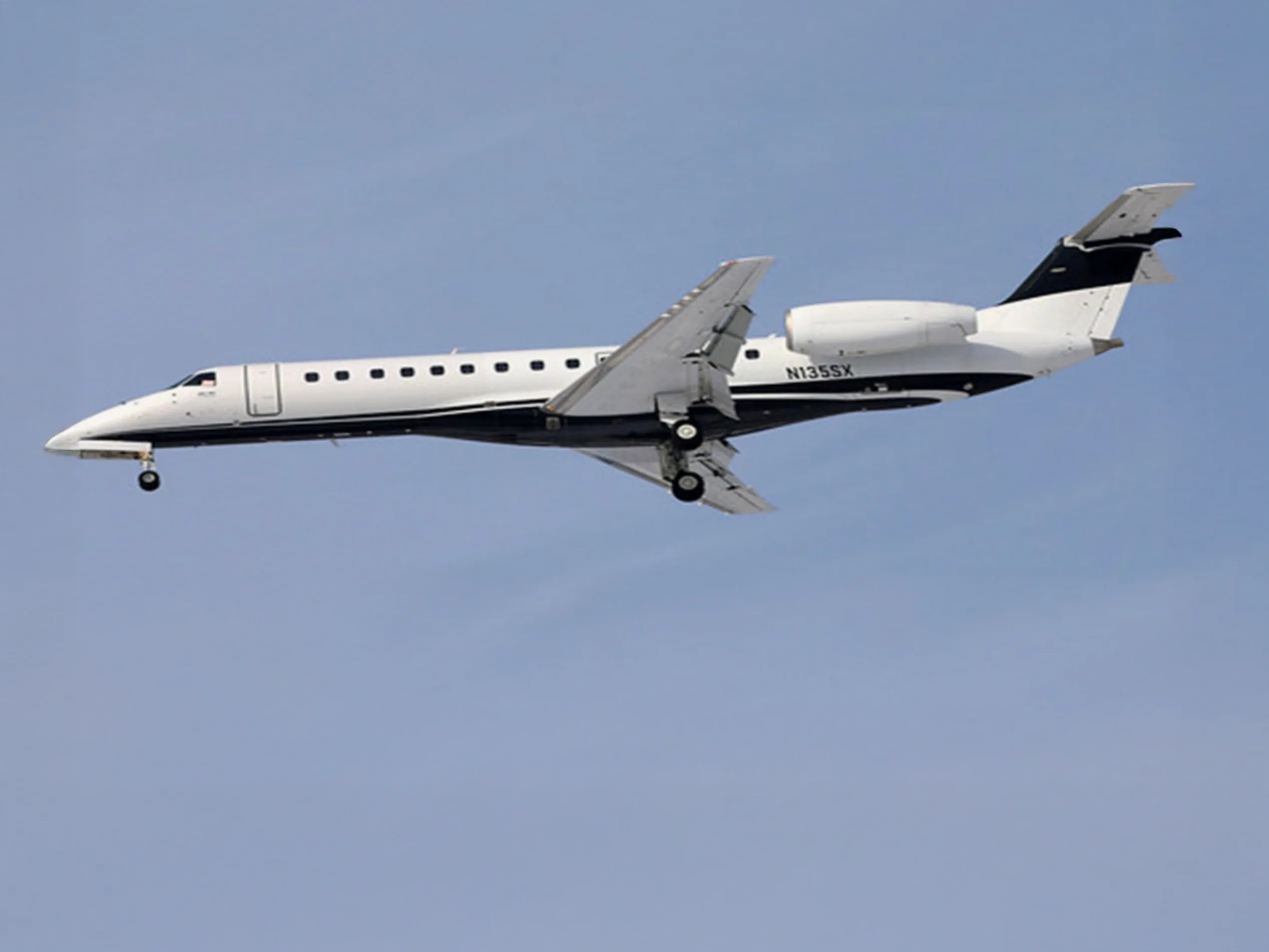 A white and black Embraer ERJ 135 aircraft with registration N135SX is seen in flight with its landing gear extended against a blue sky.