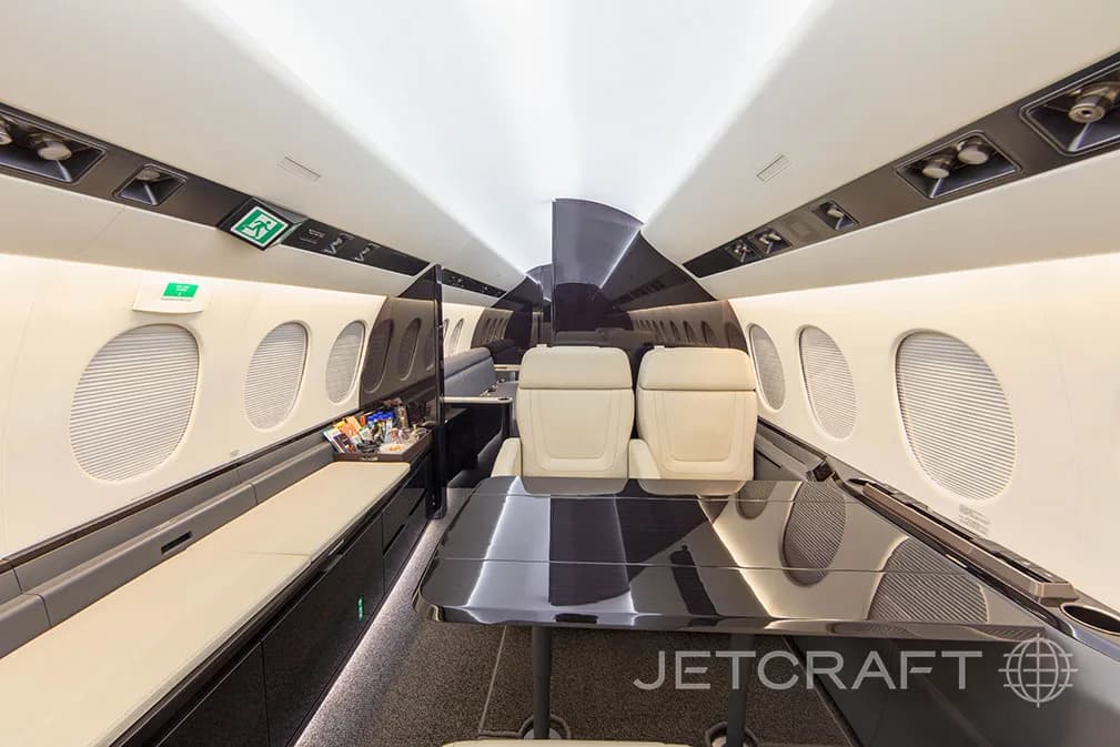A wide view of a private jet cabin, showing cream seats, dark tables, and a long side bench.