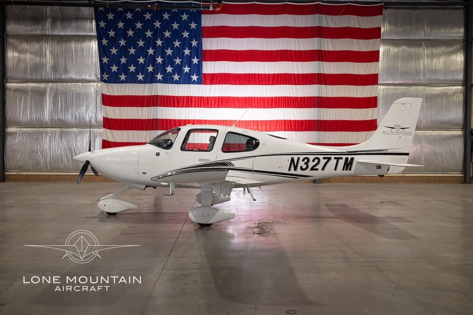 Side view of a white Cirrus SR22 aircraft with black stripes and registration N327TM, parked in a hangar with an American flag backdrop, with its gull-wing doors closed.