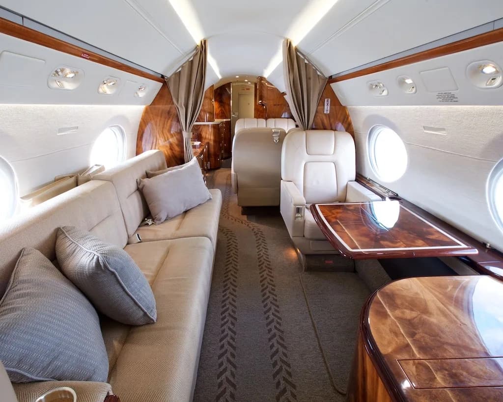 A view inside a private jet cabin showing a long beige sofa, individual seats, and polished wooden accents.