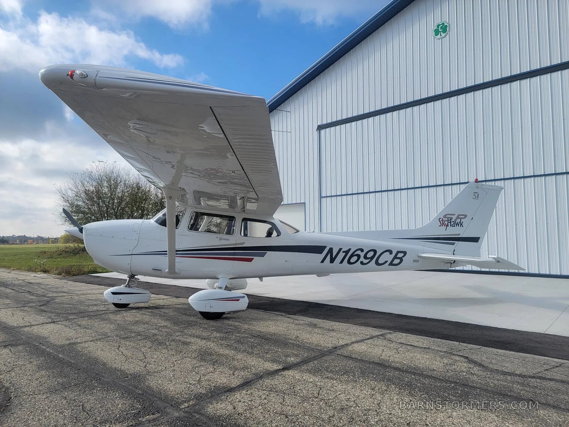 A white Cessna Skyhawk N169CB with black and red stripes is parked on an apron next to a white hangar.