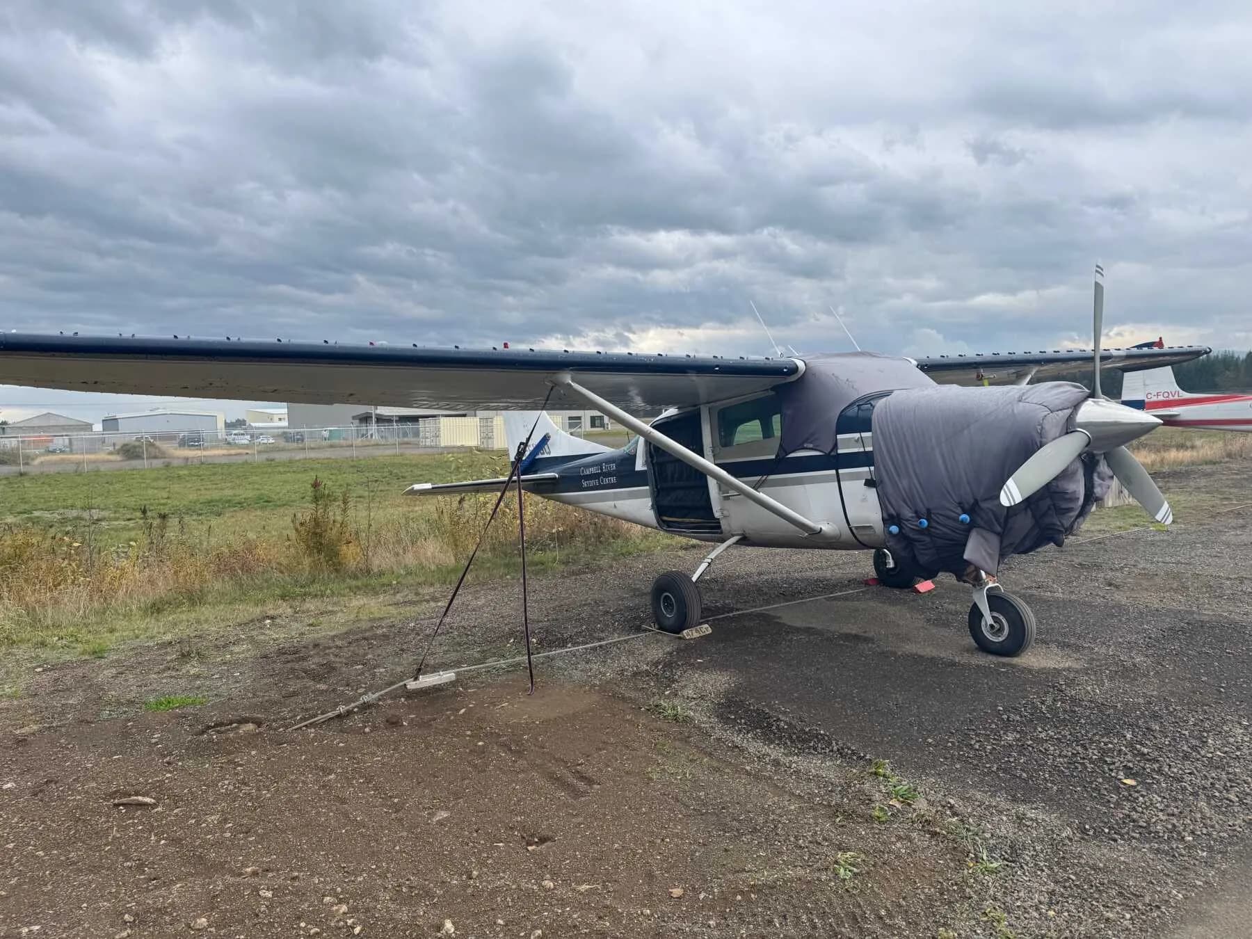 A Cessna 206 Stationair parked on a gravel area with the door open and a cover on the engine.