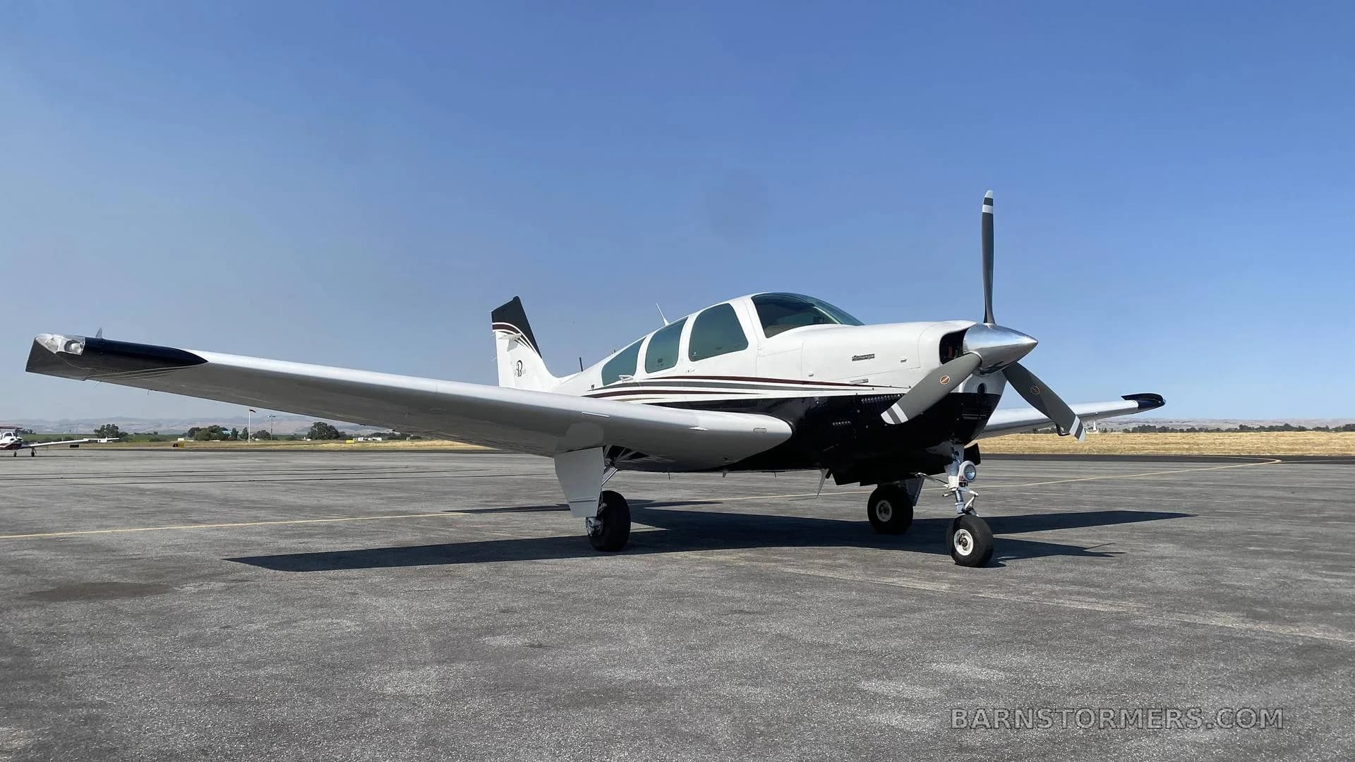 A white and black single-engine aircraft with a propeller, parked on an asphalt tarmac under a clear blue sky.