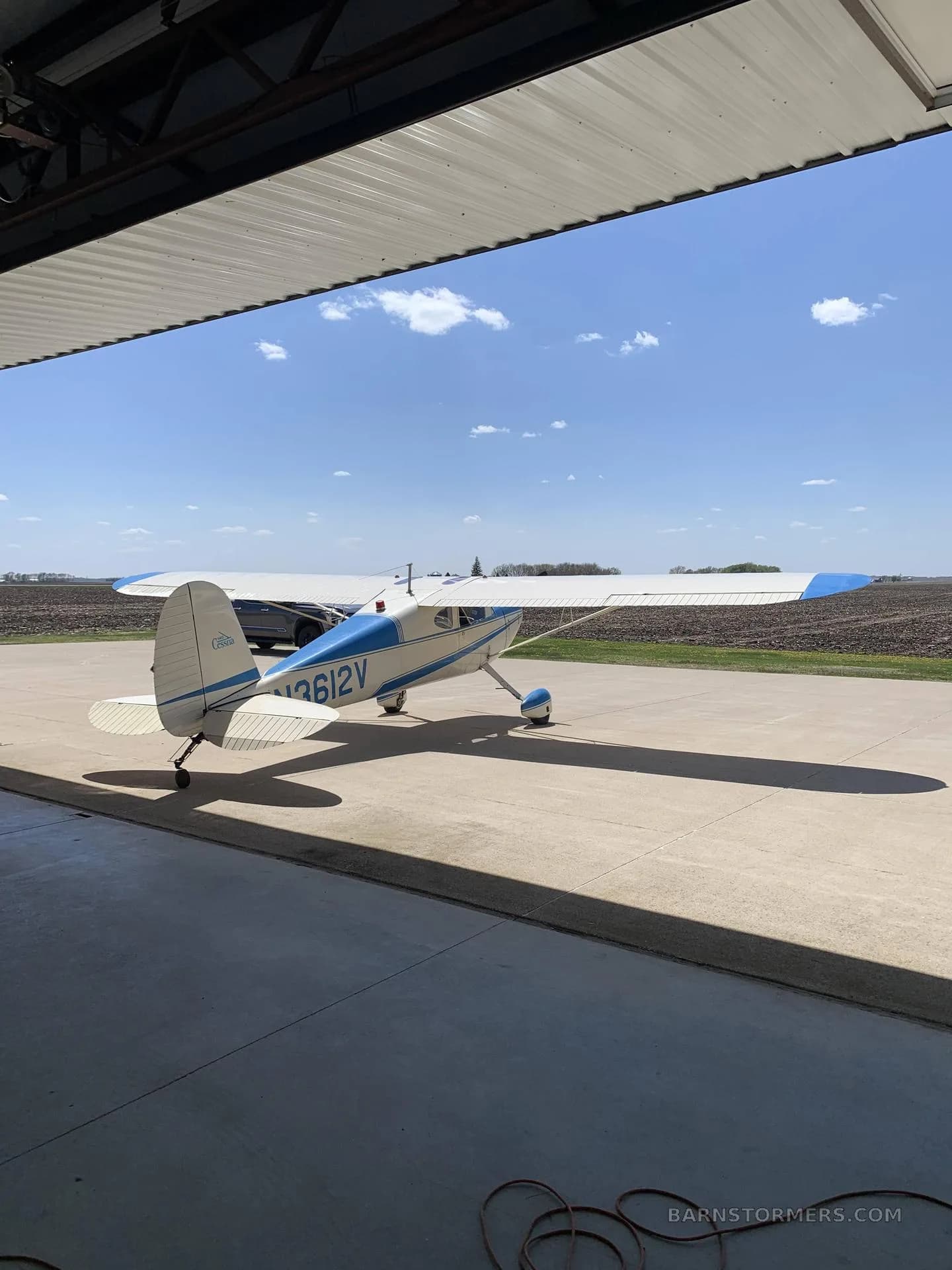 A white and blue Cessna 170B aircraft with tail number N3612V parked on a concrete pad, partially inside a hangar, with fields in the background.