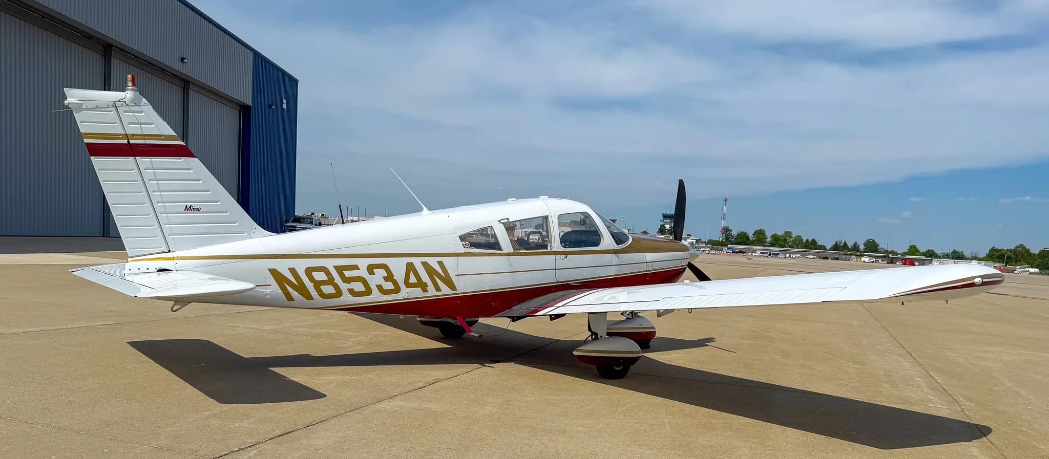 A side view of the white, red, and gold Piper Cherokee aircraft (N8534N) on an airfield under a partly cloudy sky.