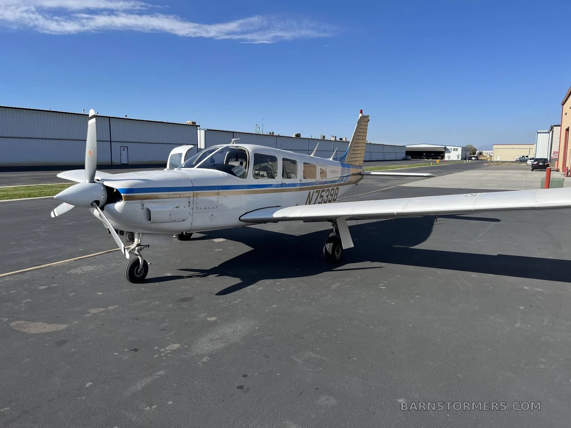 A white and blue Piper Cherokee Lance (N75398) parked on an asphalt ramp with hangars in the background under a clear blue sky.