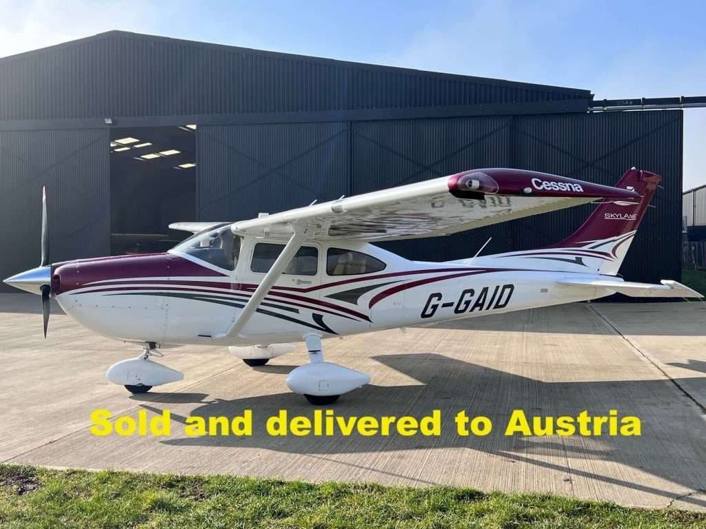 A Cessna Skylane aircraft with a maroon and white paint scheme is parked outside a hangar.