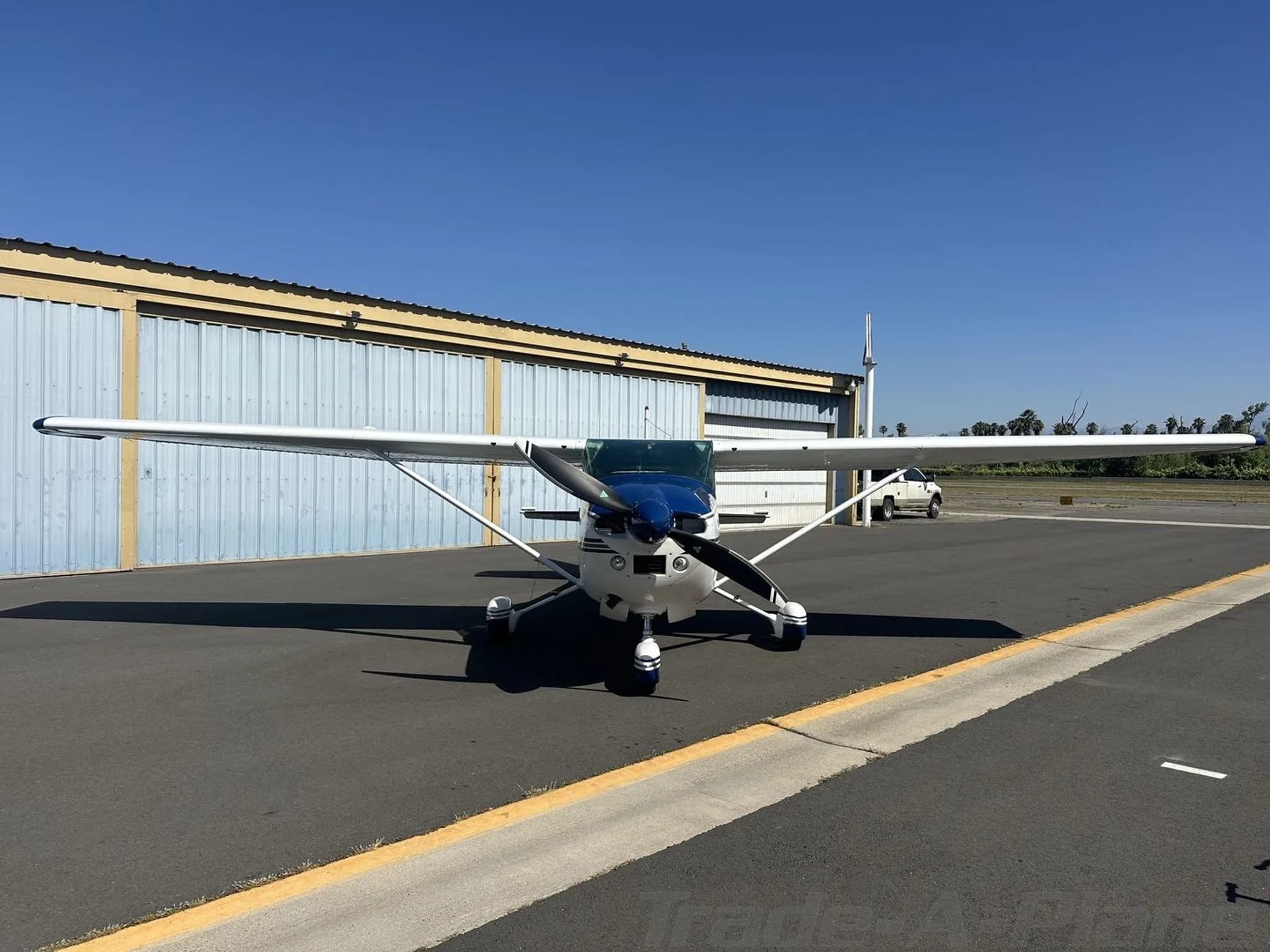 A white and blue Cessna 182 Skylane aircraft parked head-on in front of hangars.