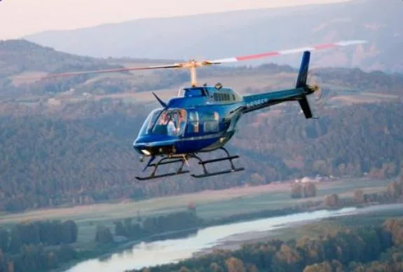 Blue Bell 206B helicopter flying over a river and forested hills.