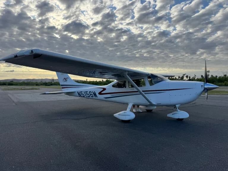 A white single-engine propeller aircraft with blue and red stripes parked on an asphalt tarmac under a cloudy sky.