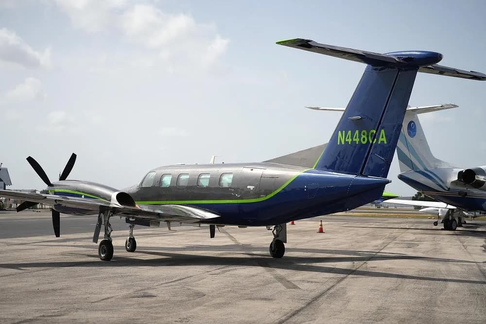 A rear-side view of a dark blue and grey twin-engine turboprop aircraft with a green stripe, tail number N448CA, on an airfield.