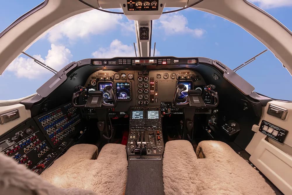 Wide view of a private jet's cockpit showing the instrument panel, controls, and a clear view of a blue sky with clouds.