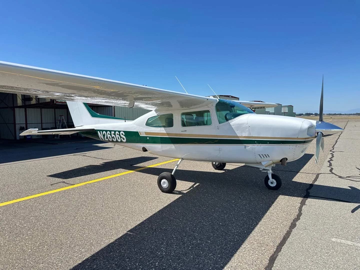 A white and green Cessna Centurion N2656S parked on an airfield, viewed from the front right with hangars in the background.