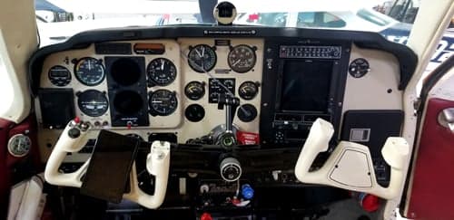 The cockpit instrument panel of an aircraft, featuring analog gauges, a digital display, and two white control yokes.
