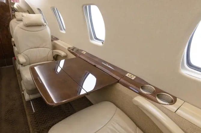 A beige leather seat next to a window in an aircraft cabin, with a polished wooden fold-out table extended.