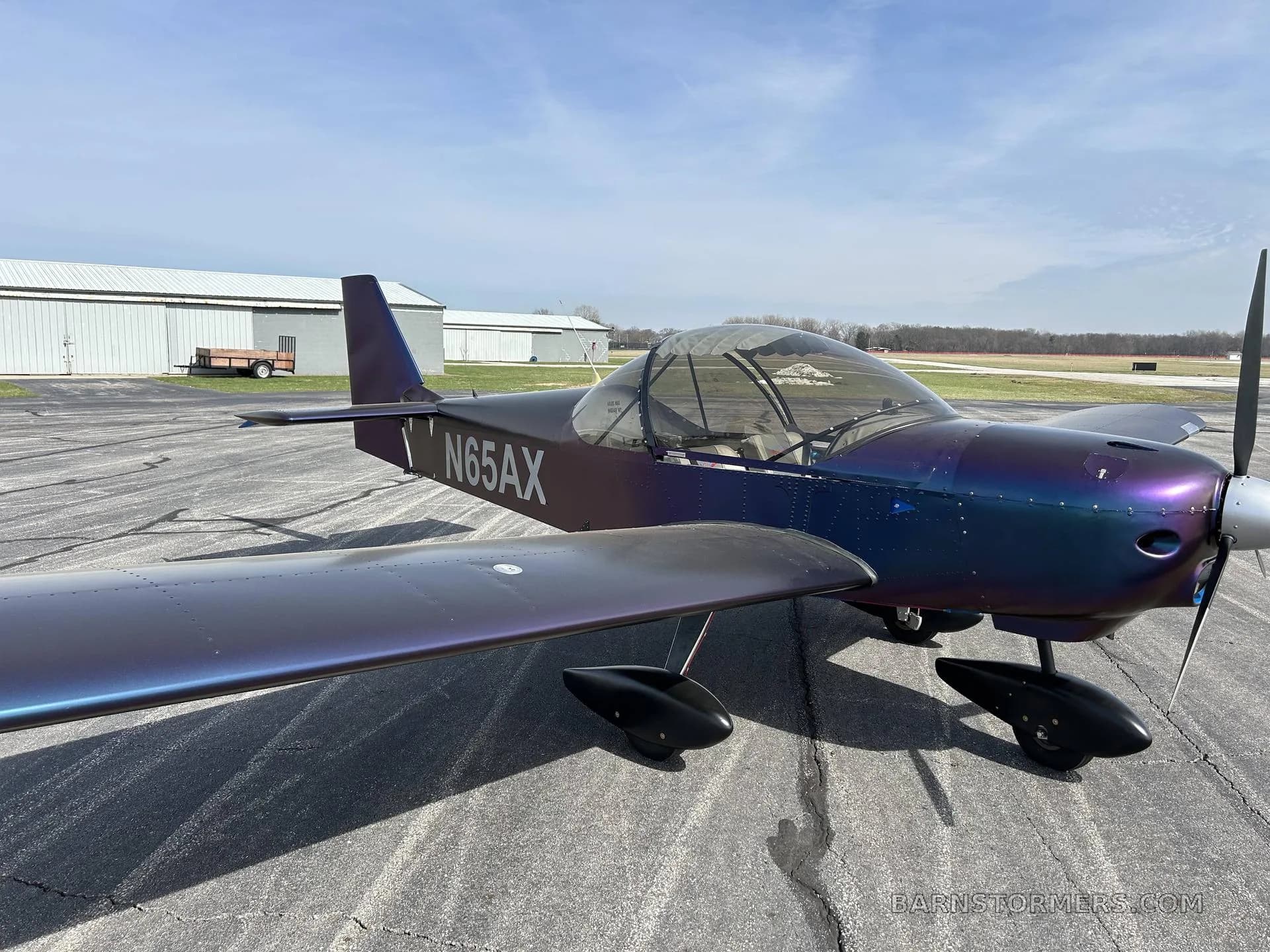 A front-right view of a purple and blue iridescent Zenith aircraft (N65AX) parked on an airport tarmac under a blue sky.