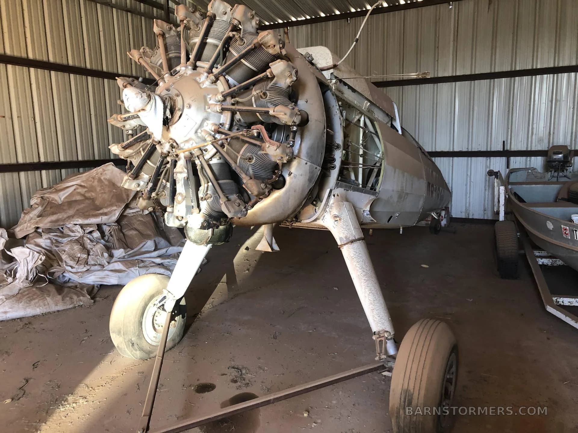 A disassembled aircraft fuselage with a large radial engine and landing gear, stored in a shed next to a boat.