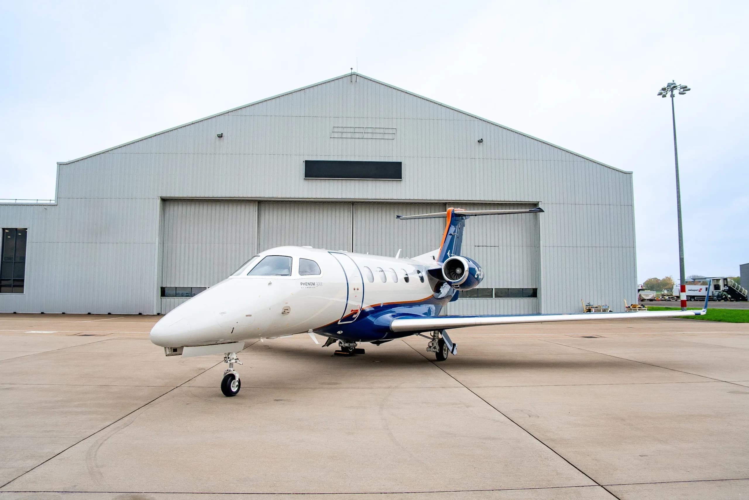 A white and blue Embraer Phenom 300E business jet with orange accents is parked on a tarmac in front of a large white hangar.
