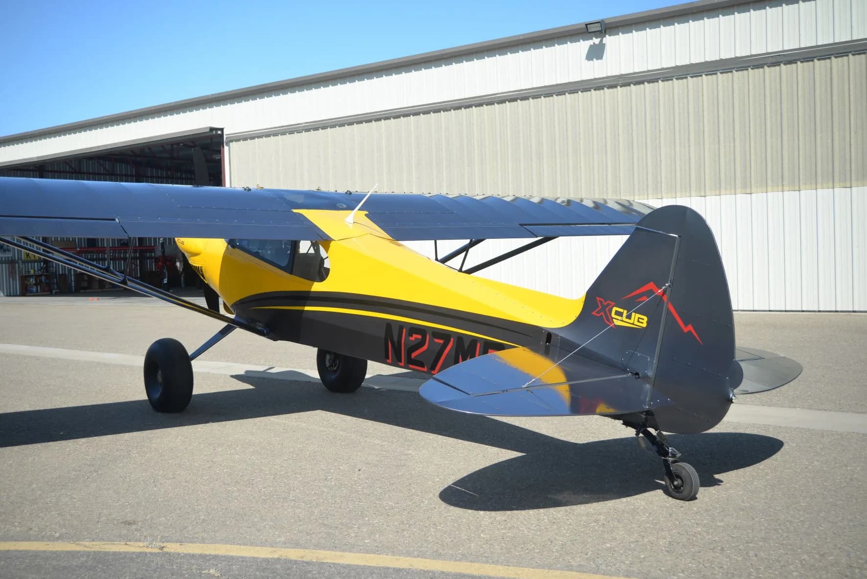 A yellow and gray CubCrafters XCub, registration N27MB, parked in front of a hangar.
