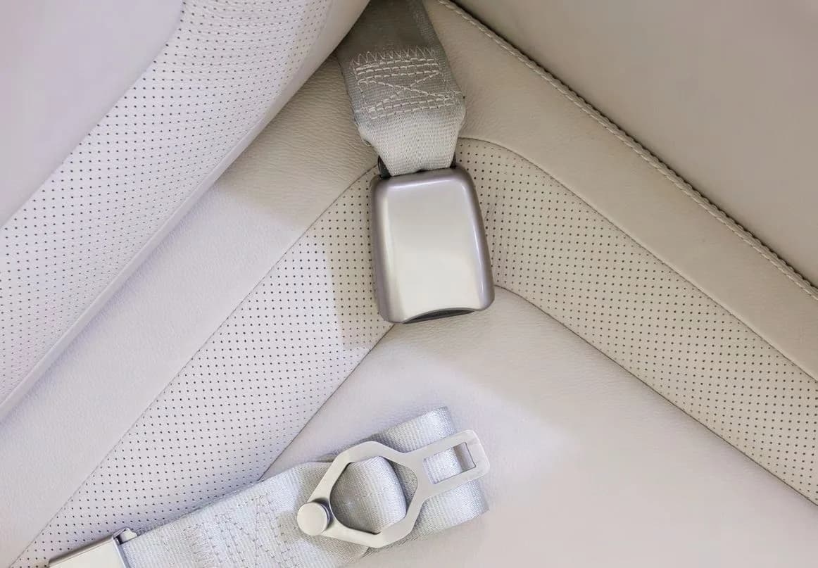 A close-up of a grey seatbelt buckle on a perforated light grey leather aircraft seat.
