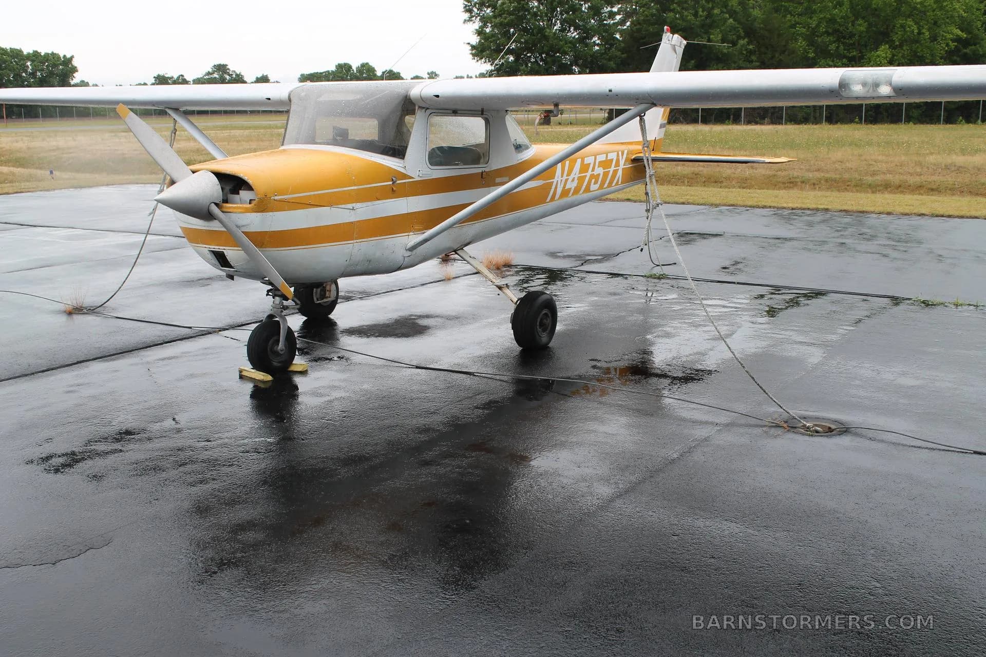 A yellow and white Cessna 150 (N4757X) is tied down on a wet tarmac, viewed from the front-left.