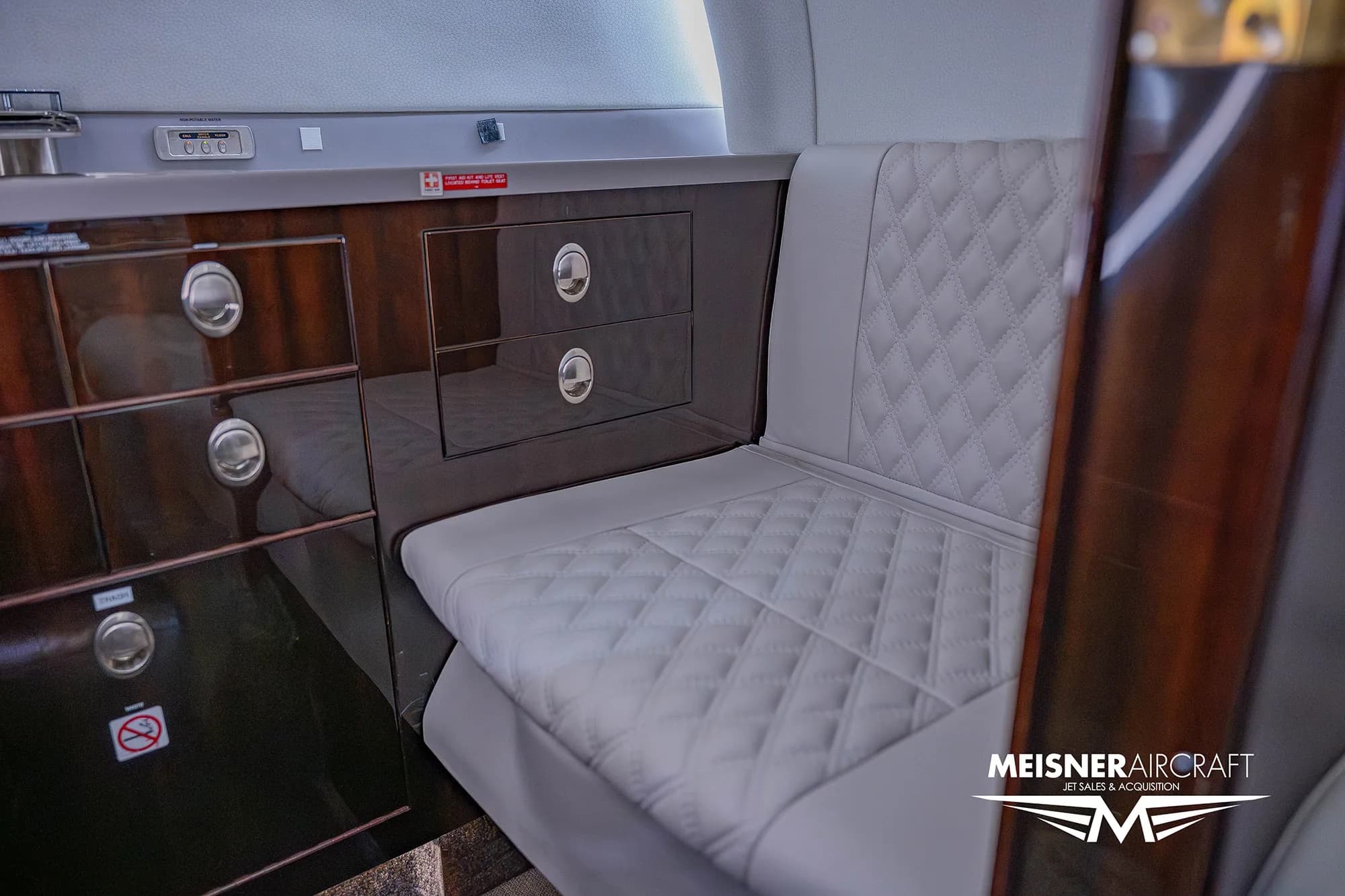 A close-up of a dark wood cabinet and a quilted white leather seat in the business jet's cabin.