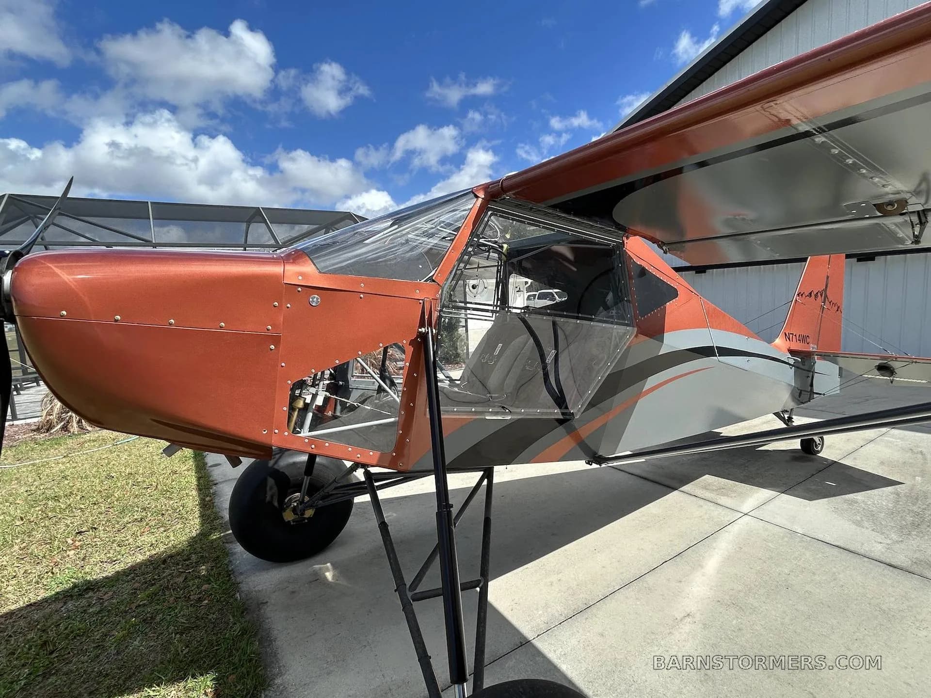 A three-quarter view of the orange and grey bush plane, showing its fuselage, wing, and open cabin door, parked next to a hangar.