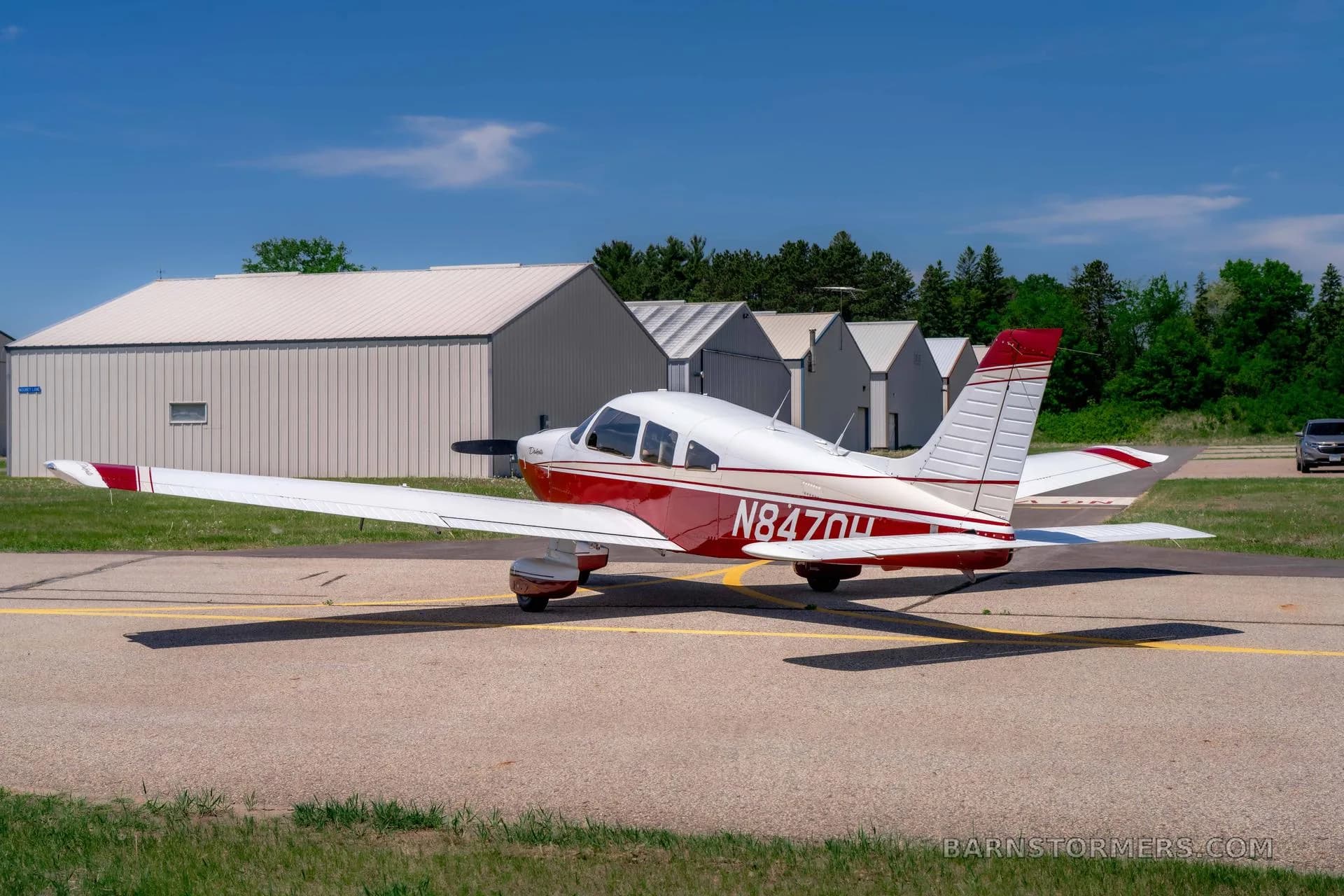 A red and white Piper Dakota aircraft with registration N847OH is parked on an asphalt tarmac, viewed from the rear quarter, with hangars in the distance.