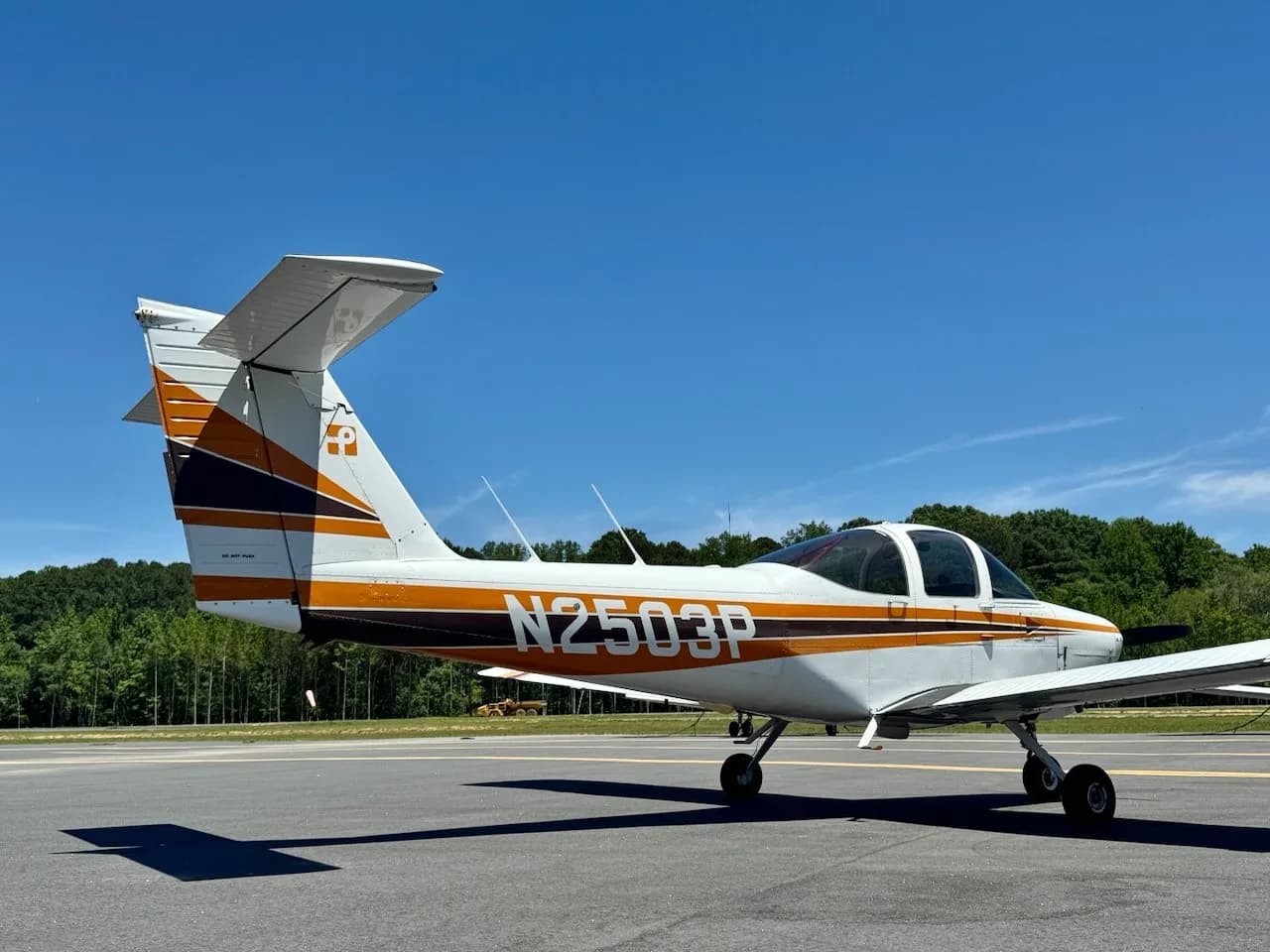 A white Piper PA-38 Tomahawk aircraft with orange and black stripes, seen from the right-rear on an airfield.