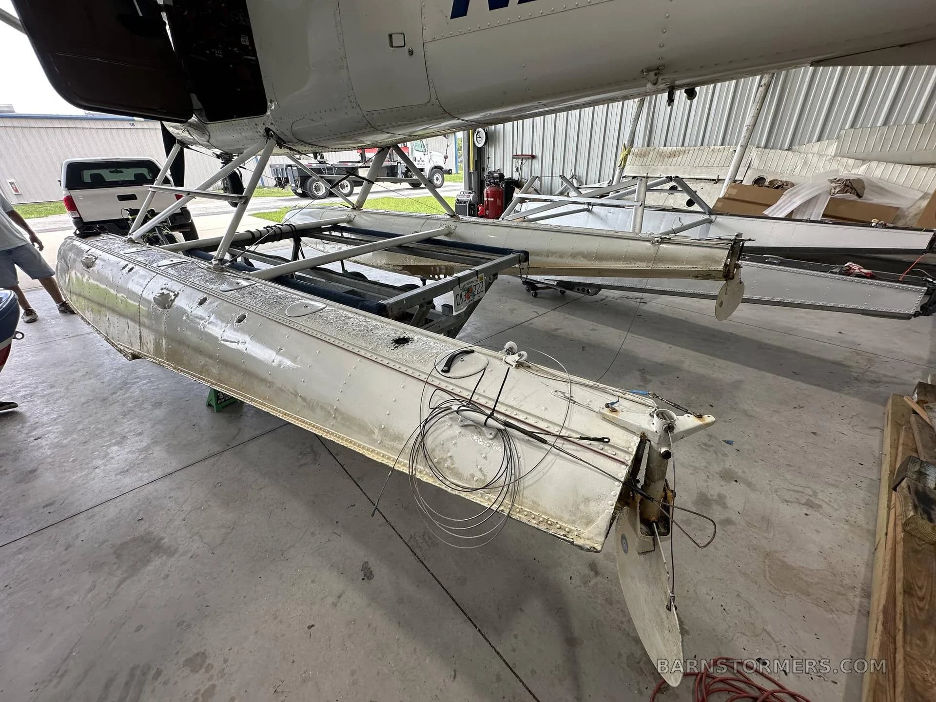 The rear section of a white floatplane's floats, with exposed wiring and control surfaces, is seen in a hangar.