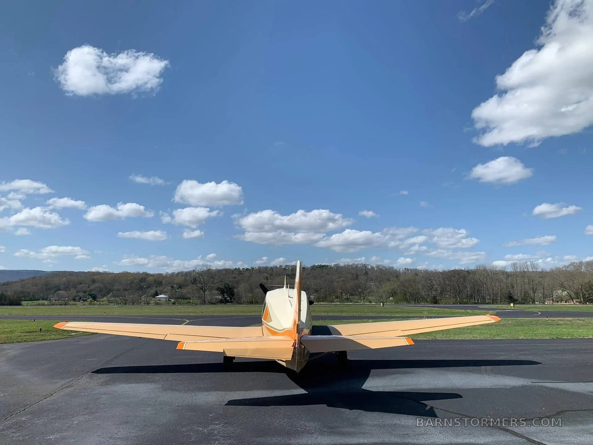 A beige and orange single-engine aircraft (N625RL) viewed from directly behind on a runway, with trees and hills in the background under a blue sky with clouds.