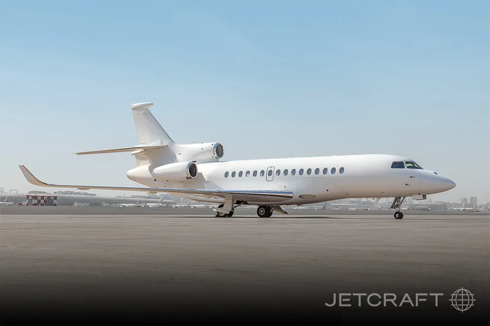 A white Dassault Falcon business jet, seen from the side on a tarmac with a distant city skyline.