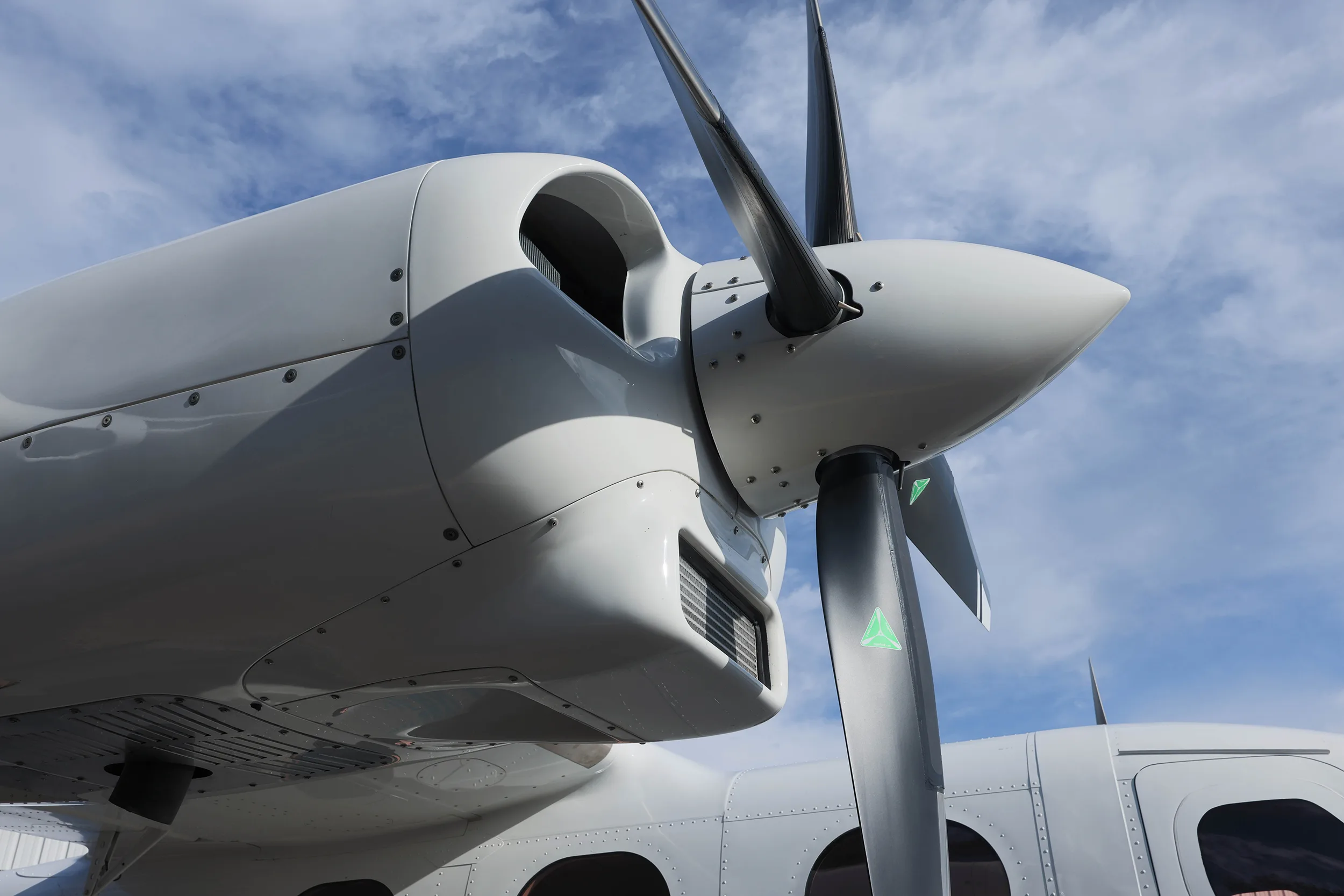 Close-up view of the white engine nacelle and three-bladed propeller of a twin-engine aircraft against a cloudy sky.