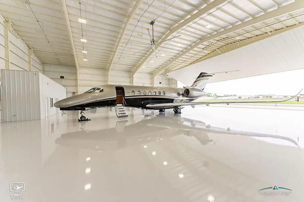 A side view of a grey and black business jet with its main door open, parked inside a large hangar.