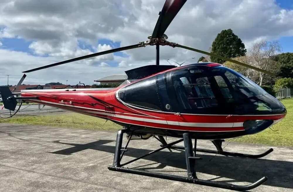 A red and black helicopter with white stripes and black skids sits on a paved surface under a cloudy sky.