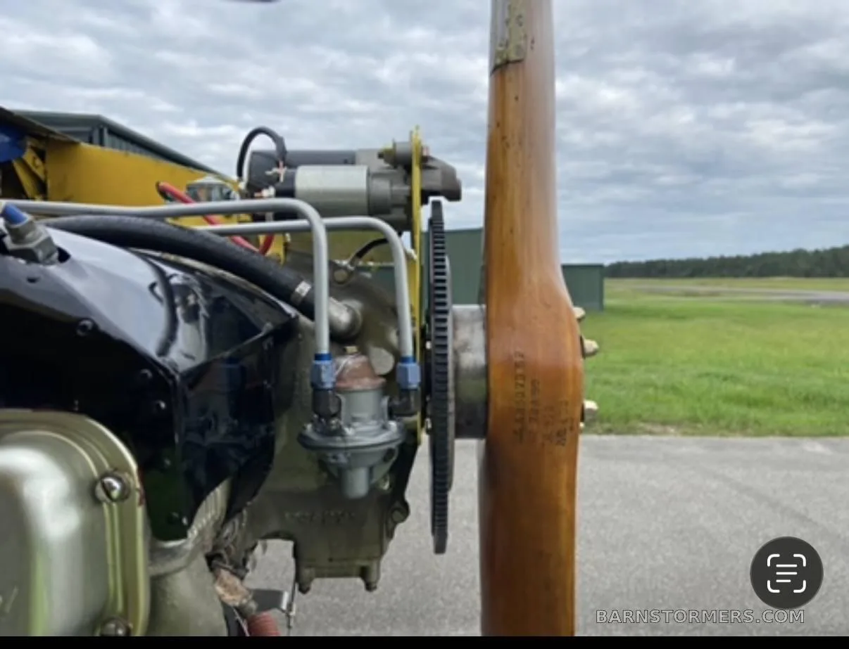 A close-up view of the engine and wooden propeller of a yellow-winged ultralight aircraft on an airfield.