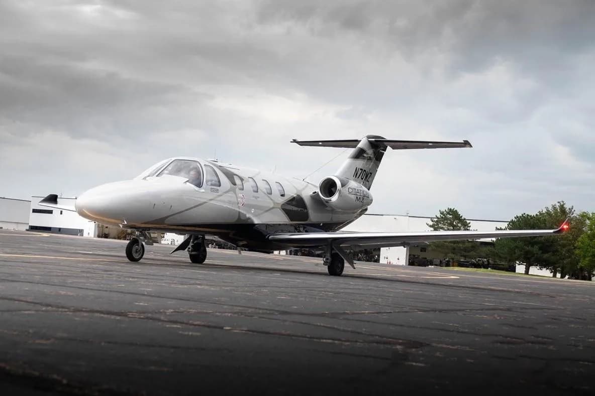 A low-angle front-right view of a silver and black Cessna Citation M2 jet (N70MZ) on an asphalt ramp under a cloudy sky.
