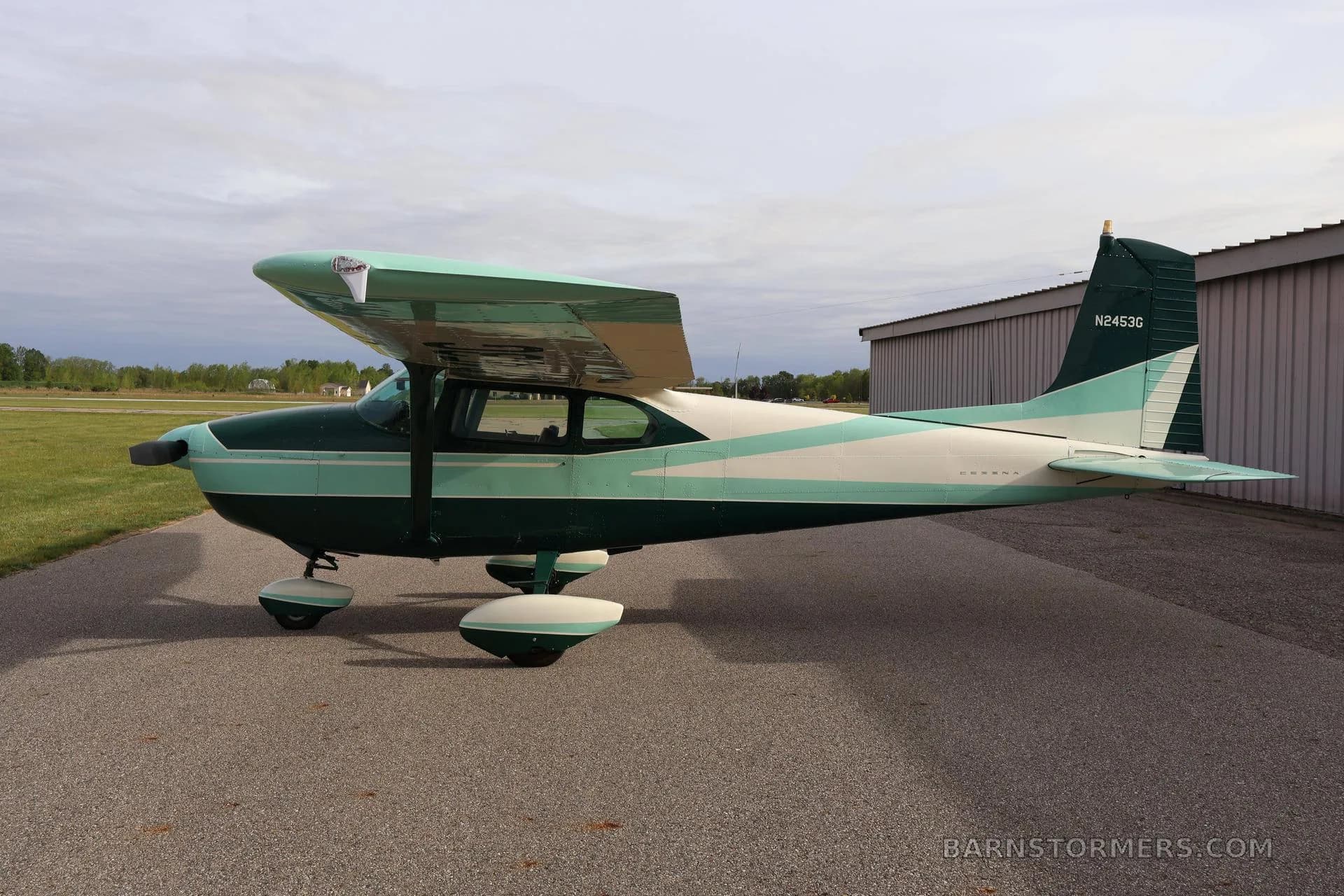 A green and white single-engine propeller aircraft (Cessna 172) is parked on an asphalt ramp next to a hangar, viewed from the side.