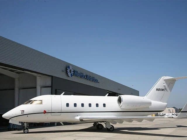 A white Bombardier Challenger business jet parked on an airport tarmac in front of a "Western Aviation" hangar.