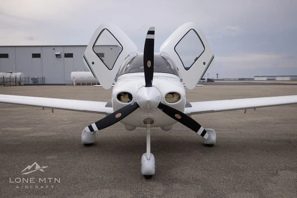 A head-on view of a white Cirrus SR22 aircraft with its propeller and open doors visible, parked on an asphalt ramp.