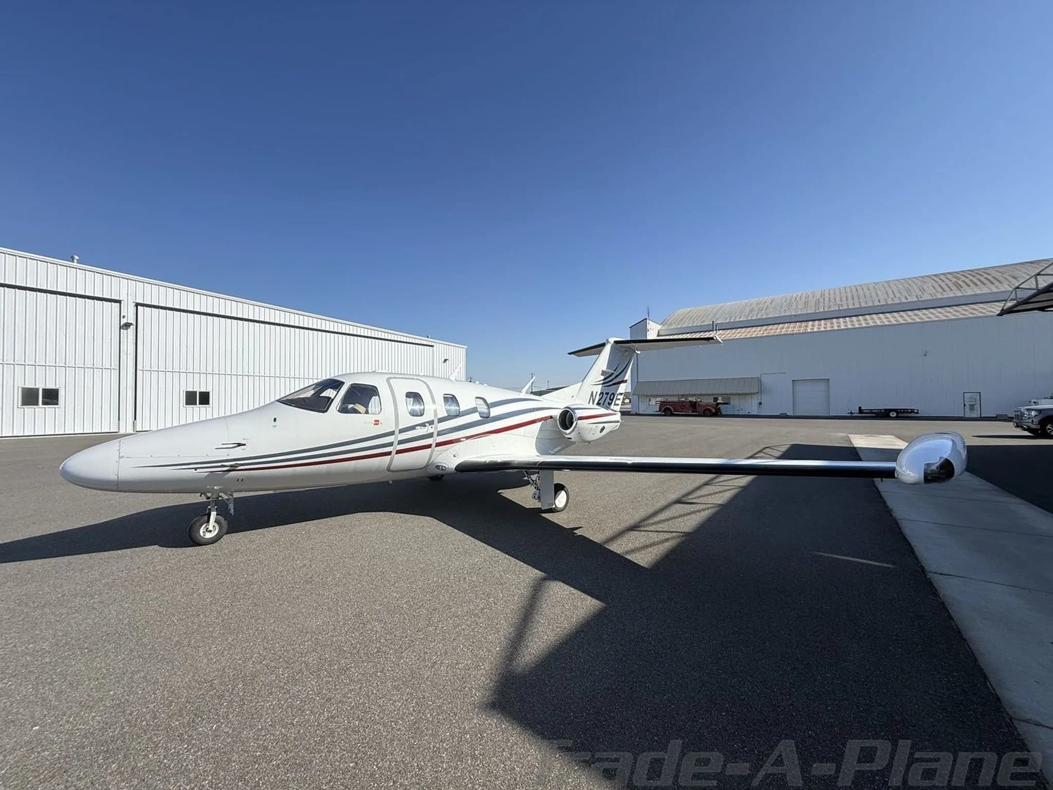 Side view of a white private jet with red and blue stripes and registration N279E parked on an apron.