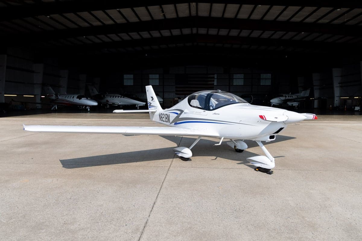 A white Tecnam Astore light sport aircraft with blue and grey stripes parked on a tarmac in front of an open hangar.