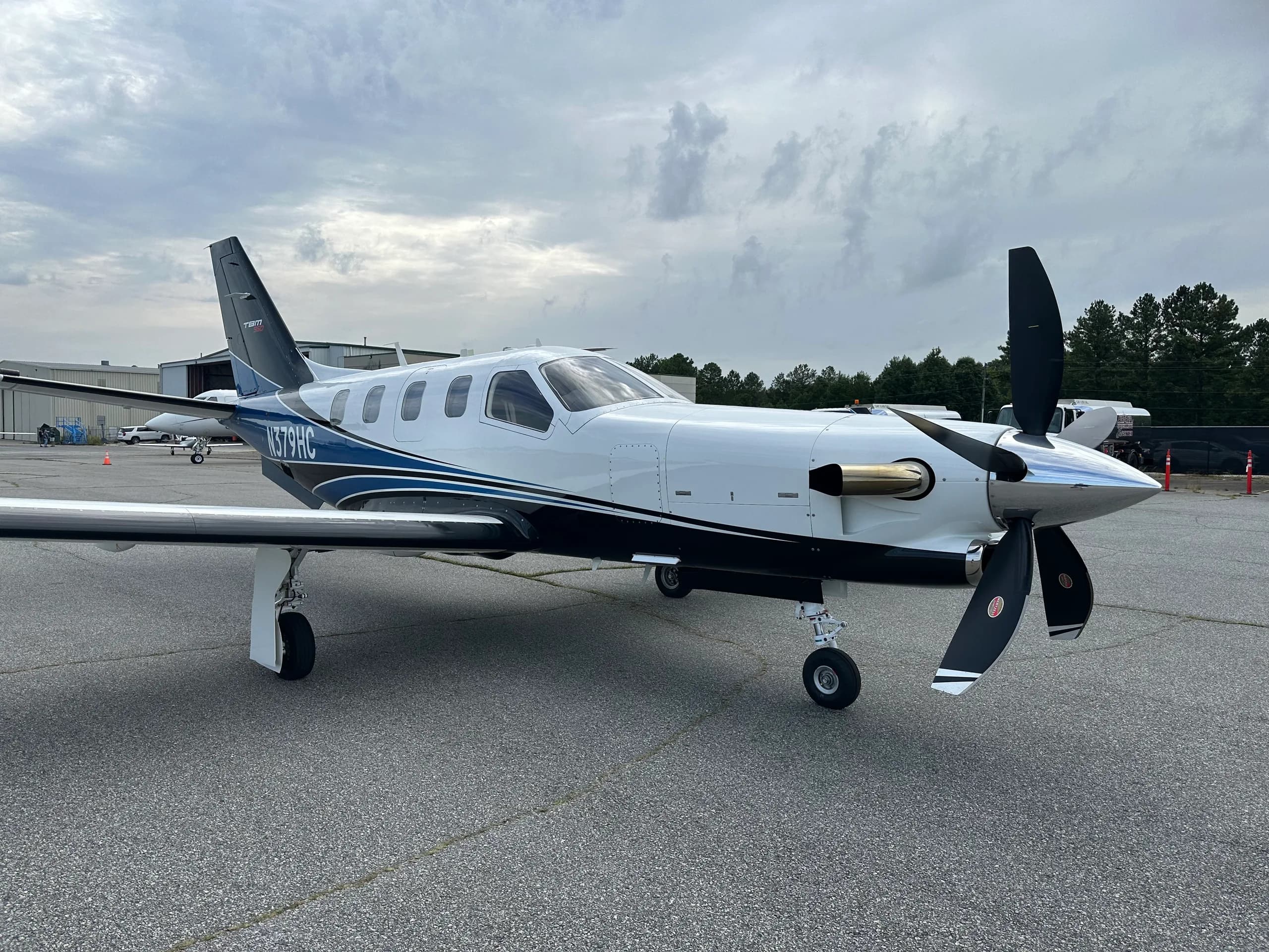 A white and blue TBM 960 turboprop aircraft with a black propeller, parked on an asphalt tarmac under a cloudy sky.