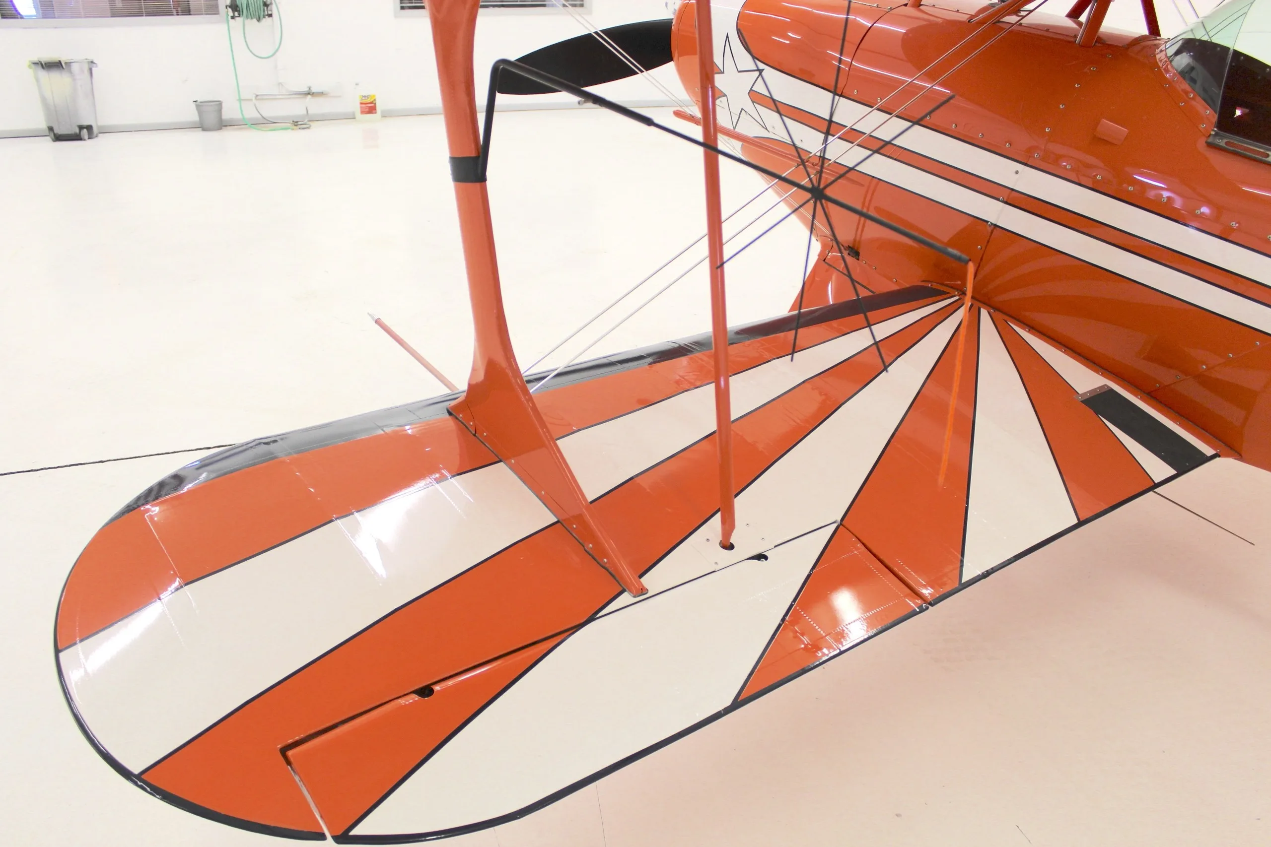 An overhead close-up view of the upper wing of an orange Pitts S-1T biplane, featuring a white and orange sunburst pattern.