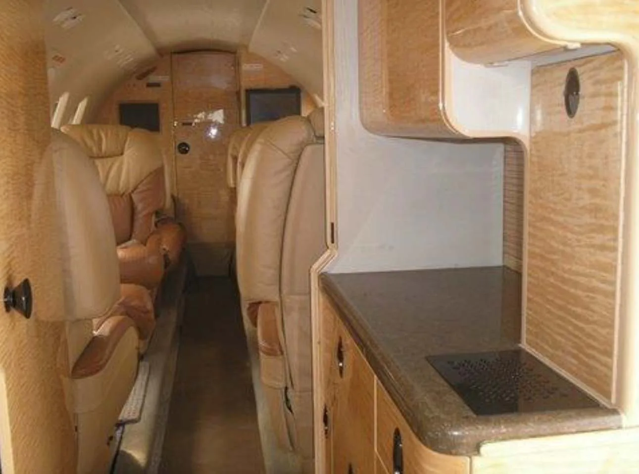 The interior cabin of a private jet, showing a wooden galley area with a counter and seats further down the aisle.