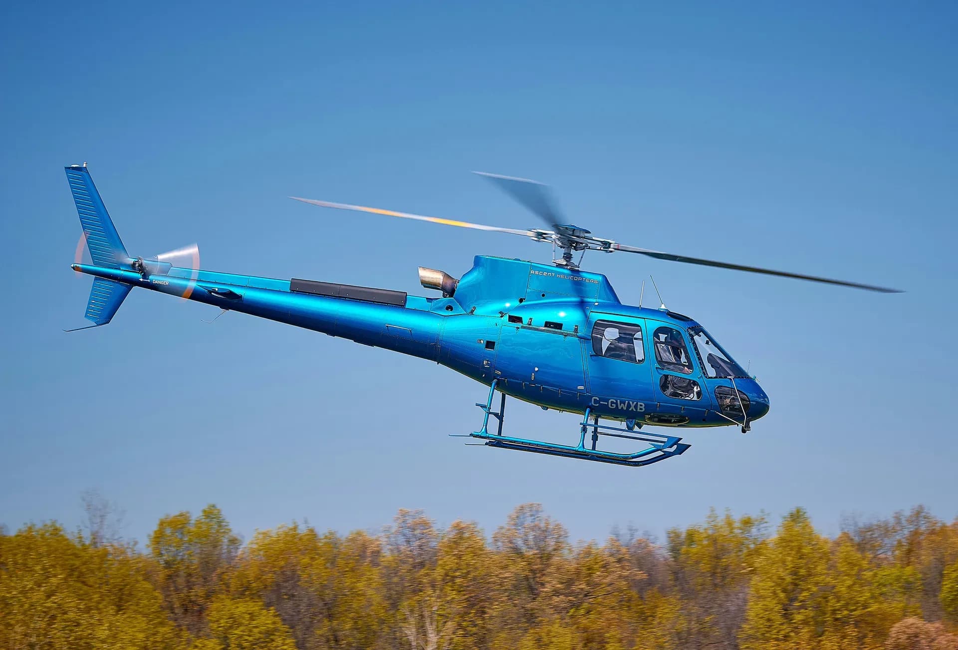A blue Airbus H125 helicopter with registration C-GWXB flying against a clear blue sky.
