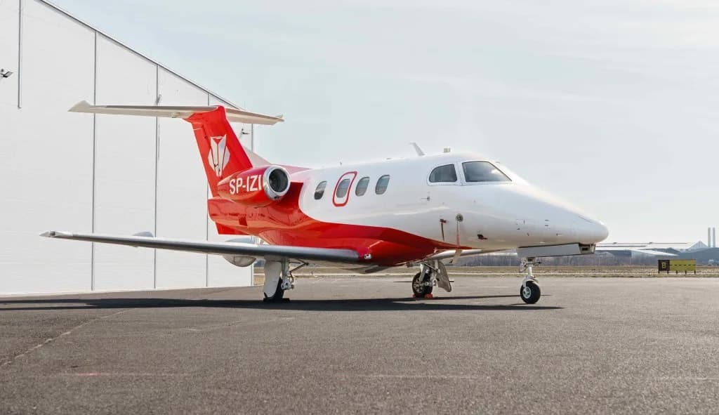 A white and red Embraer Phenom 300, registration SP-IZI, parked on an airport tarmac next to a hangar.
