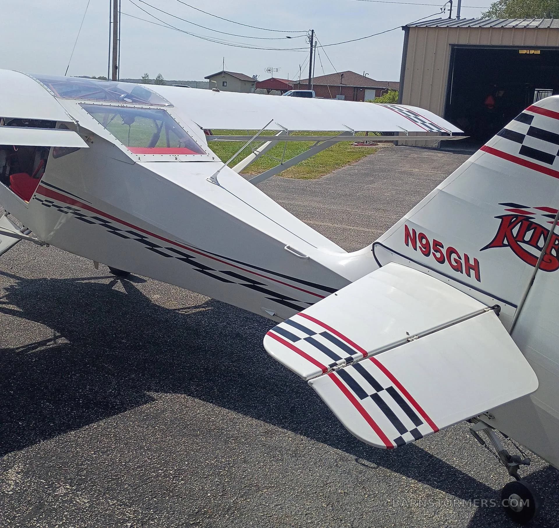 The tail section of a white SkyStar Kitfox aircraft with red and black checkerboard stripes is shown outdoors.